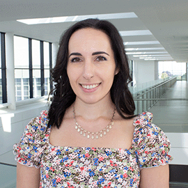 A smiling woman stands in front of a university corridor as she looks directly ahead.